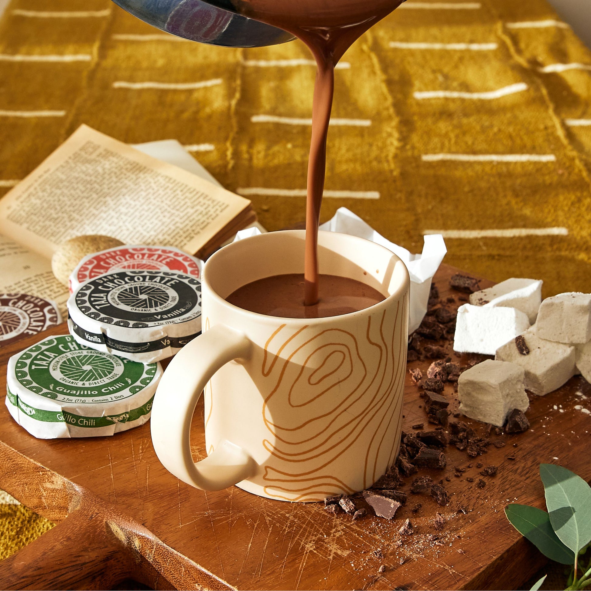 Hot Chocolate being poured into a mug placed near Taza Chocolate Discs and scattered ingredients.