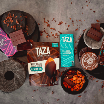 Taza dark chocolate bars, discs and chocolate covered nuts on a textured surface with various Taza packaging and ingredients.