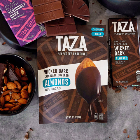 Bag of Wicked Dark Chocolate Covered Almonds on a stone background surrounded by exposed chocolate and almonds.